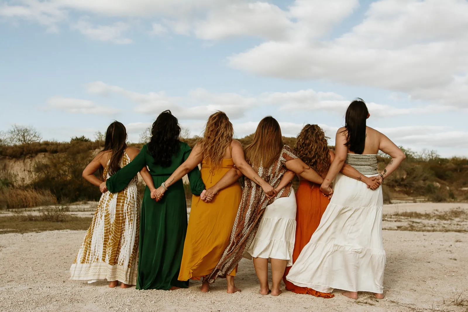 Women in flowing dresses with arms around each other, facing the landscape